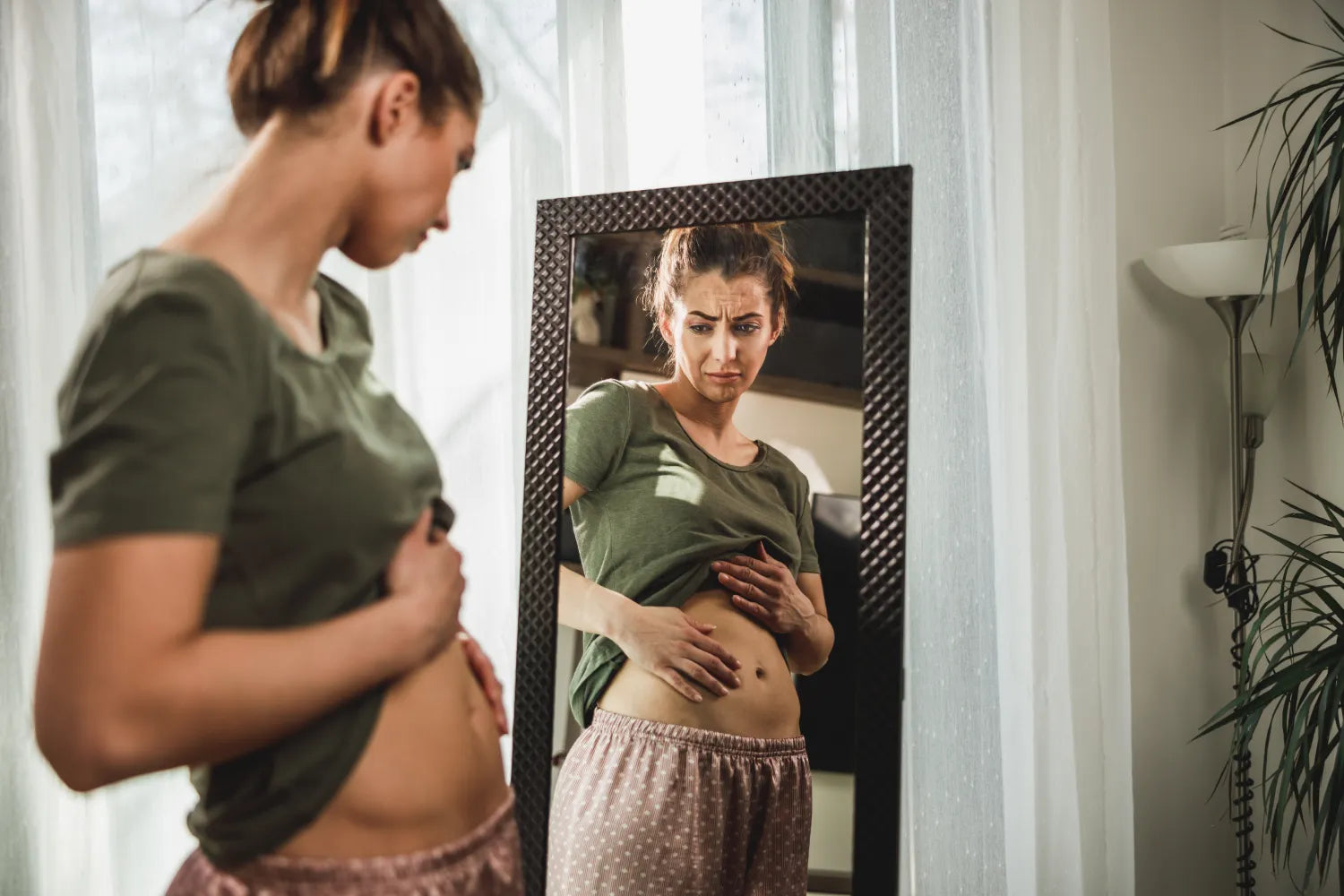 Woman looking concerned as she examines her bloated stomach in a mirror in a bedroom