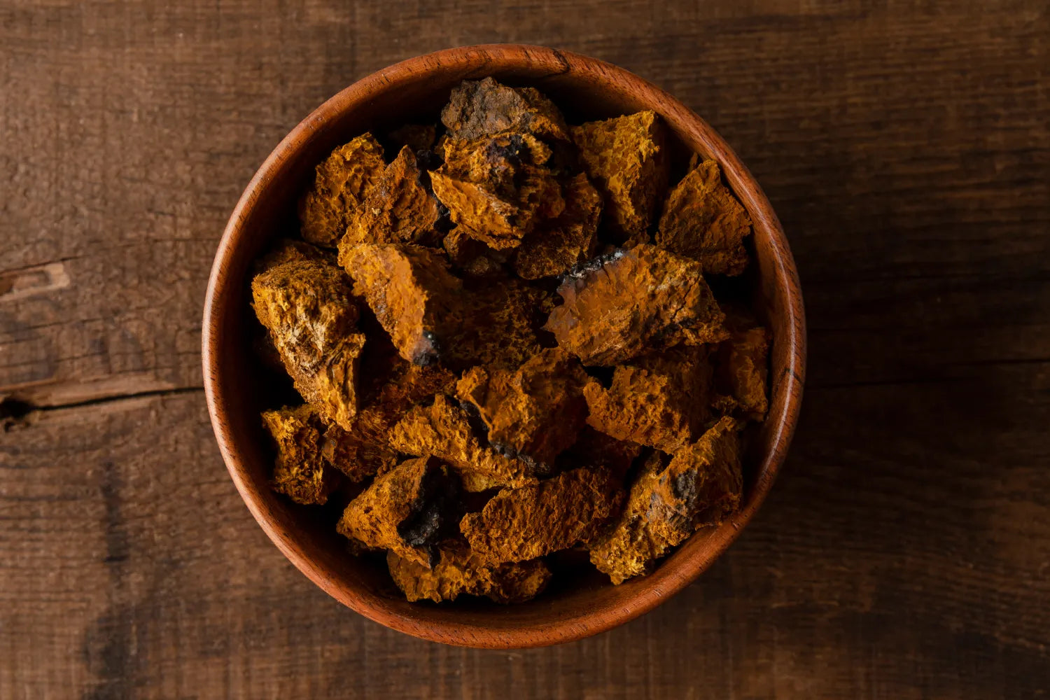 Top-down view of dried chaga mushroom chunks in a wooden bowl showing rough orange-brown porous texture on dark wooden table
