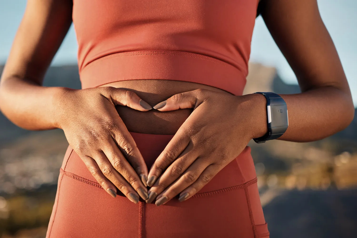 Person making a heart with hands on their stomach wearing a rust workout set and a fitness tracker outdoors