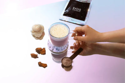 Hand holding a frothy mushroom coffee latte in a clear mug with Everyday Dose packet, powder scoop, and dried mushrooms