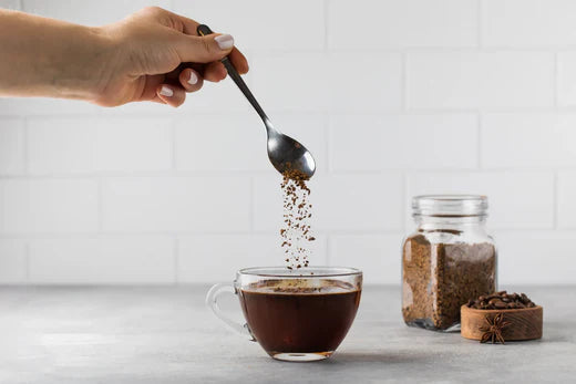 Hand sprinkling instant coffee into a glass cup, jar and bowl of coffee on a gray countertop