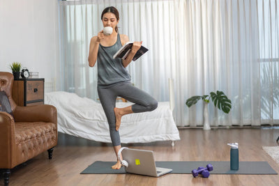 Woman balancing on one leg drinking coffee in a bedroom with yoga mat, laptop, dumbbells and water bottle