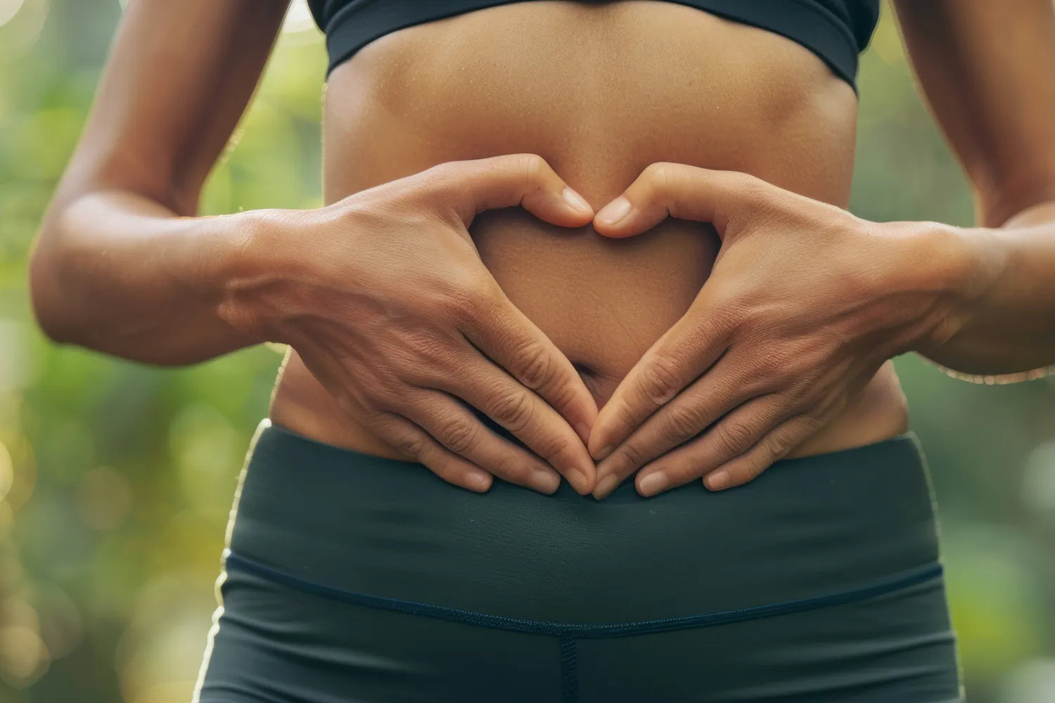 Close-up of a person forming a heart with their hands over their bare abdomen to symbolize gut health, blurred outdoor background