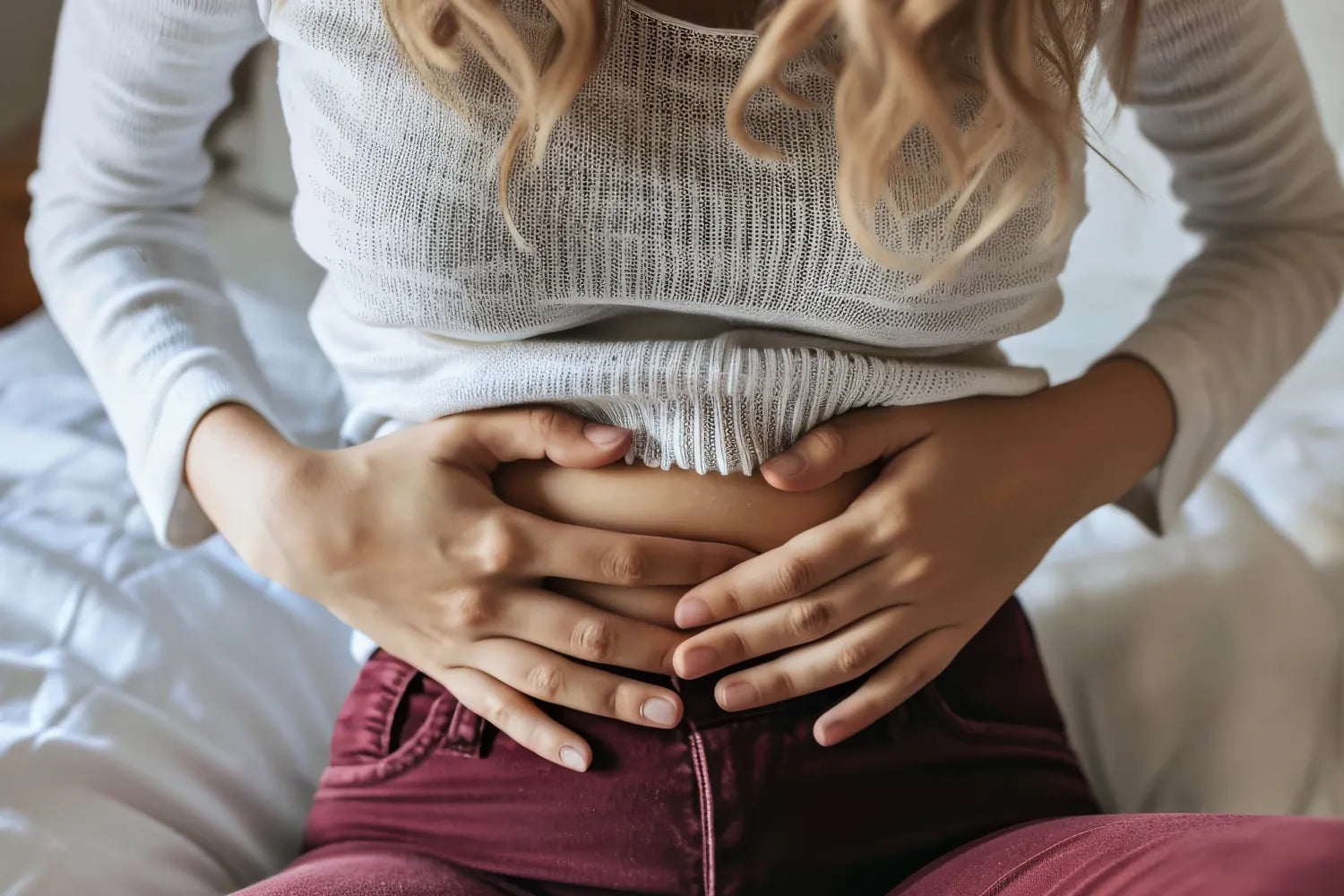 Woman sitting on a bed holding her lower abdomen in discomfort, wearing a white knit sweater and maroon pants