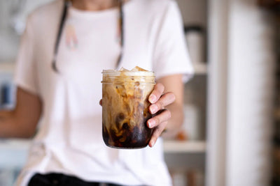 Person holding a mason jar of iced breve with cream swirling into espresso in a bright cafe setting
