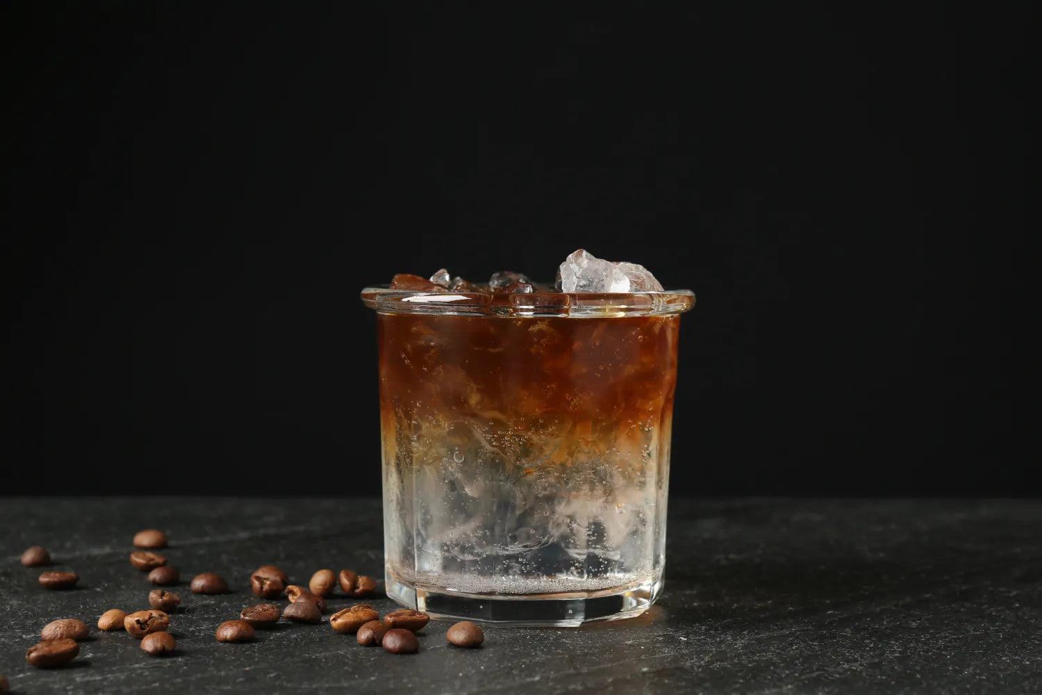 Cold brew cocktail in a short glass over ice on a black background with scattered coffee beans