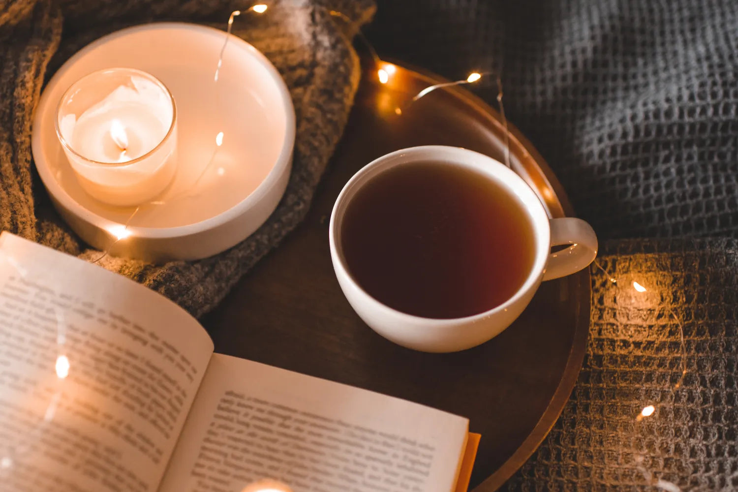Cup of tea beside an open book and lit candle on a knit blanket, cozy reading scene with warm string lights