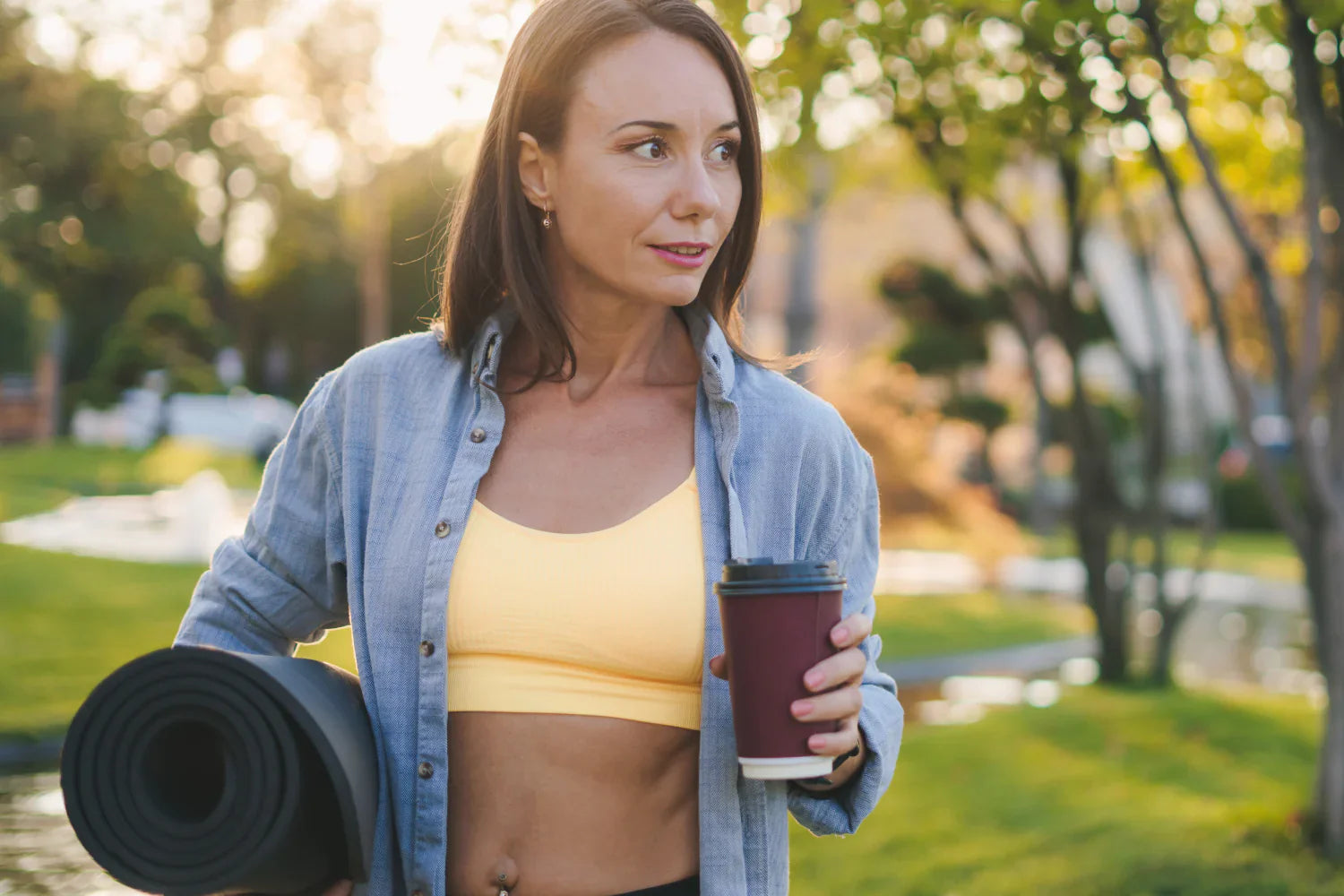 Woman carrying a rolled yoga mat and holding a takeaway coffee cup in a sunlit park after a workout