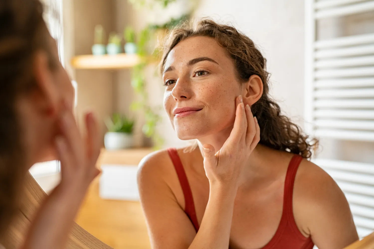 Woman touching her cheek and smiling at her reflection in a sunlit bathroom
