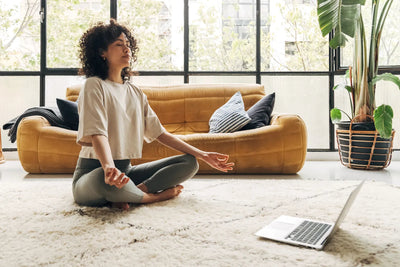 Woman meditating cross-legged on a rug in a bright living room with a yellow sofa and a laptop nearby