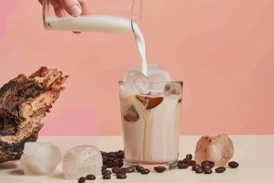 Front view of iced mushroom coffee showcasing milk being poured over ice with scattered coffee beans and a piece of mushroom on a pink background