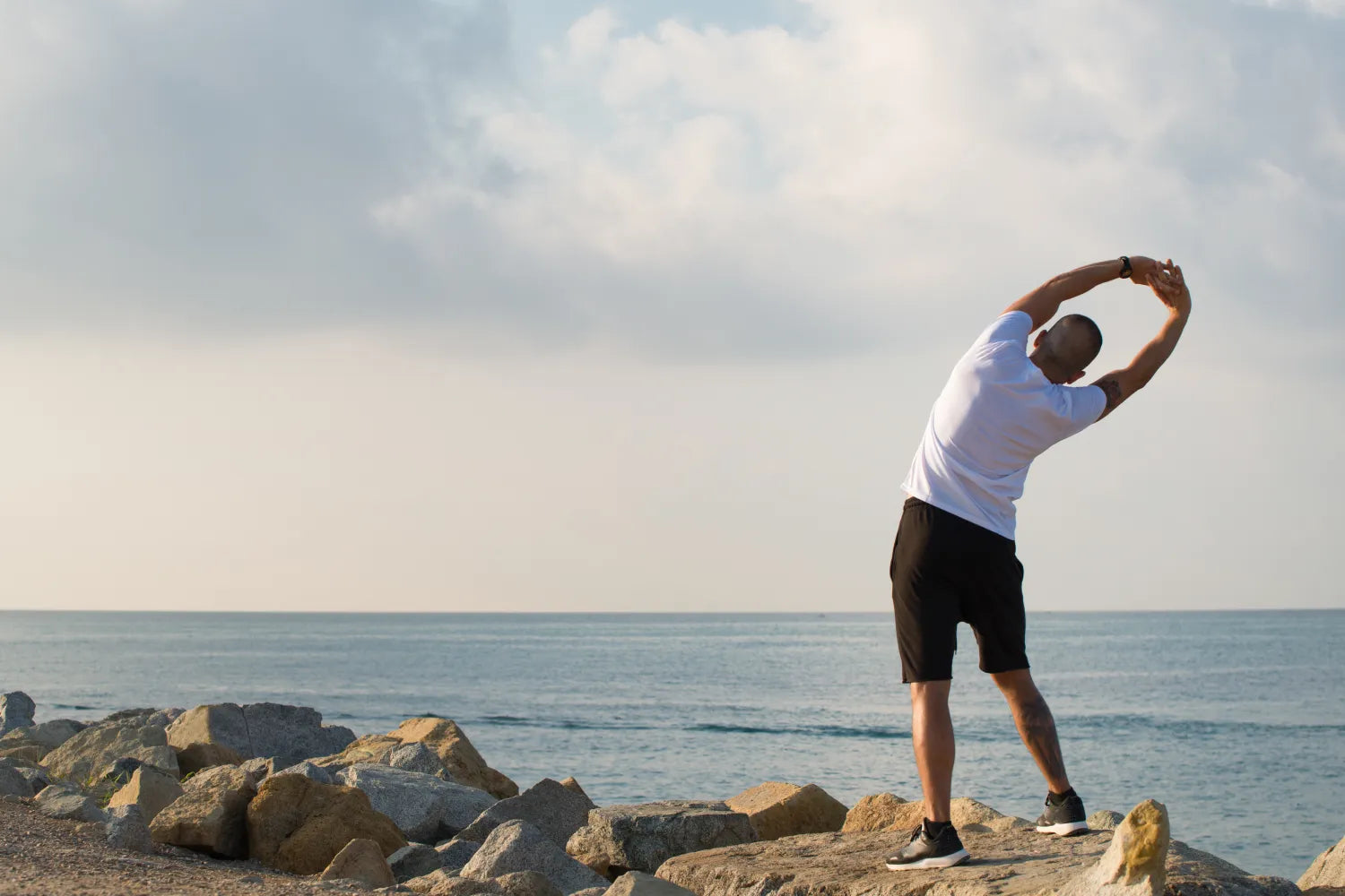 Man standing on rocks by the ocean stretching his arms overhead in a side bend against a cloudy sky
