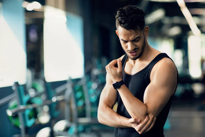 Man in a black tank top holding his elbow in pain at a gym with blurred weight racks behind him