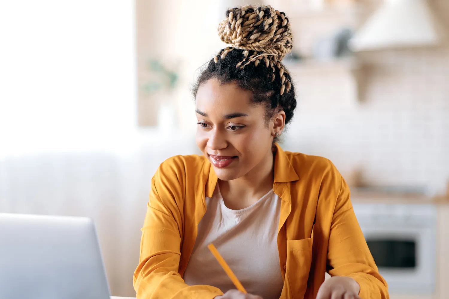 Young woman taking notes while working on a laptop in a bright home kitchen