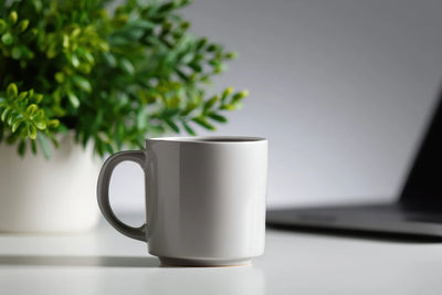 White ceramic mug on a desk with a blurred potted plant and laptop in the background
