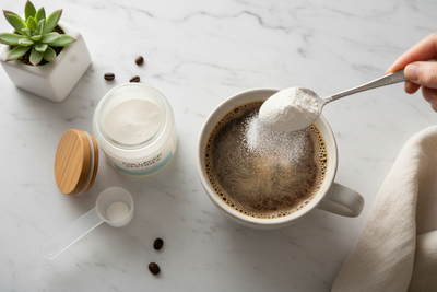 Hand sprinkling collagen powder into a mug of hot coffee on a marble countertop with an open jar and scoop nearby