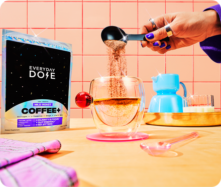 Angled view of Everyday Dose Coffee Plus bag with a hand pouring coffee powder from a measuring spoon into a double-walled glass, pink tiled wall and blue tumbler in the background