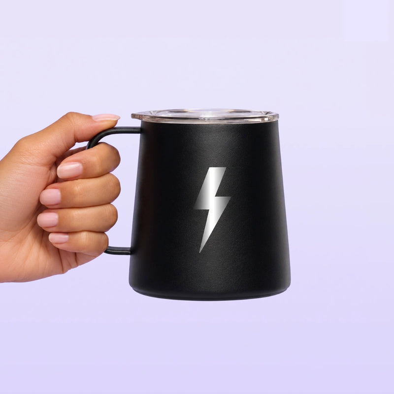 Black travel mug with a handle, clear lid, and white lightning bolt emblem held by a hand against a lavender background