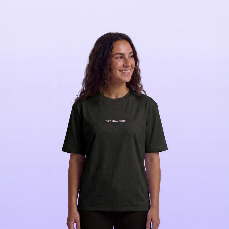 Smiling woman wearing a dark charcoal Everyday Dose T shirt with a small chest logo against a soft lavender background