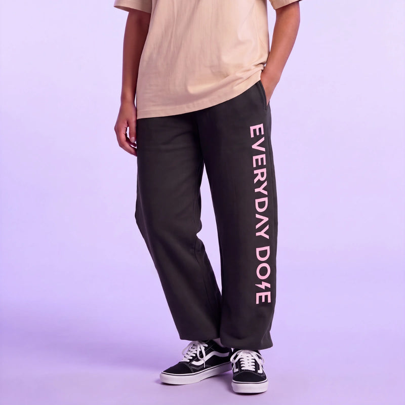 Model wearing black sweatpants with pink vertical Everyday Dose lettering a cream shirt and black sneakers against a lavender background