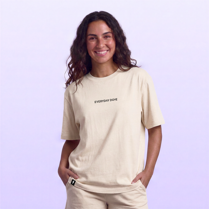 Smiling woman wearing a relaxed beige Everyday Dose T shirt with matching sweatpants against a soft lavender background