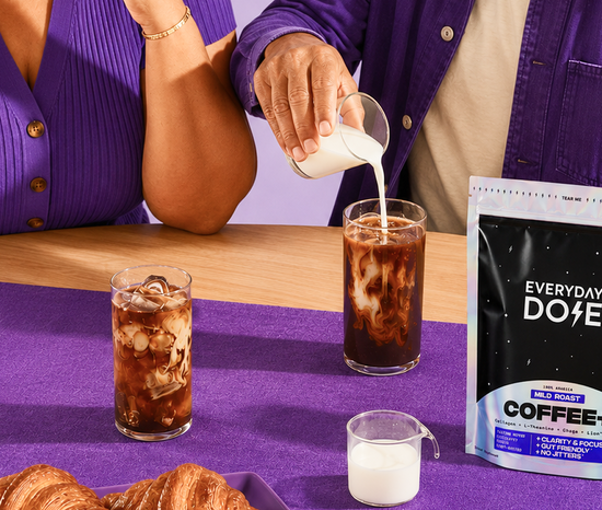 Two glasses of iced Everyday Dose coffee with milk being poured, Everyday Dose coffee bag and croissants on a purple tablecloth