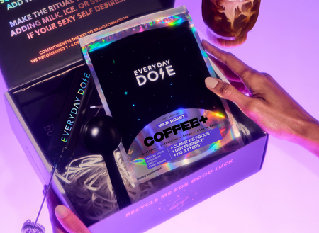 Hands holding an open box containing a holographic packet labeled Coffee+ with a black scoop inside on a purple surface
